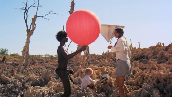 un niño aborigen de piel oscura con un globo rojo en sus manos junto a dos niños blancos.